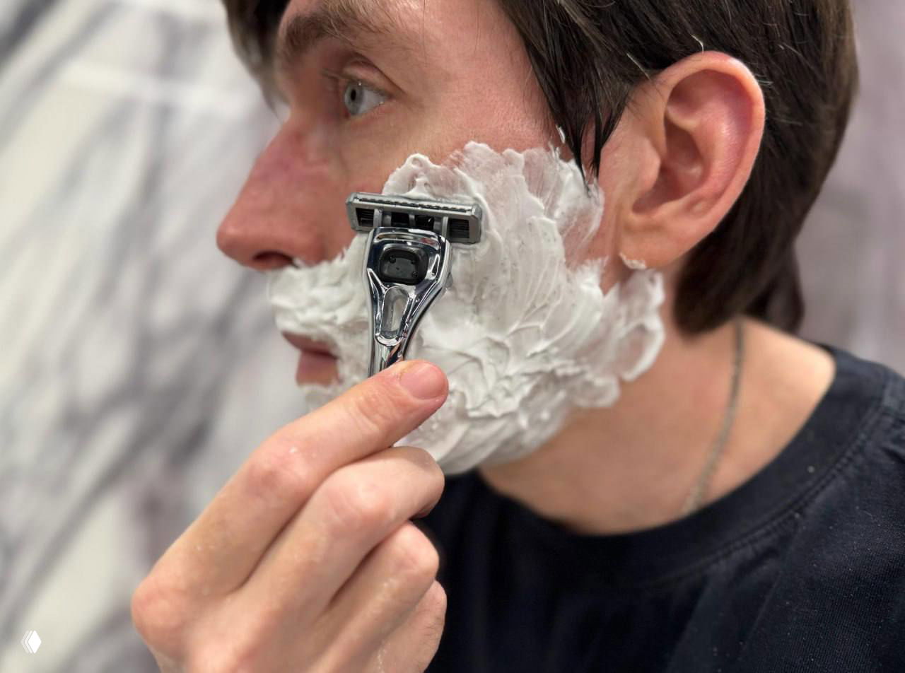 Крупный план: лезвие бритвы Damsler на пенистой щеке мужчины; демонстрация прилегания лезвия и контакта с кожей.