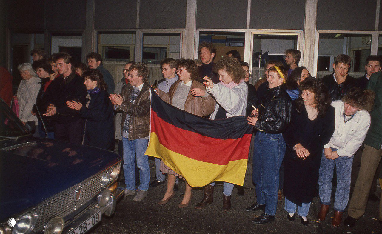 Группа людей держит флаг Германии и стоит у подъезда, выражая радость и надежду; сцена отражает национальные символы и общественную реакцию на открытия границ.
