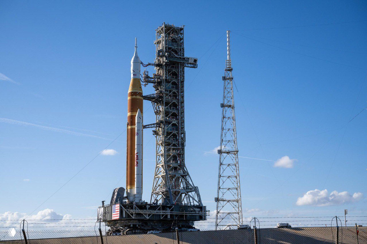 Ракета SLS установлена на пусковой платформе рядом с обслуживающей мачтой, вид с фона ясного неба и ограждений территории старта.