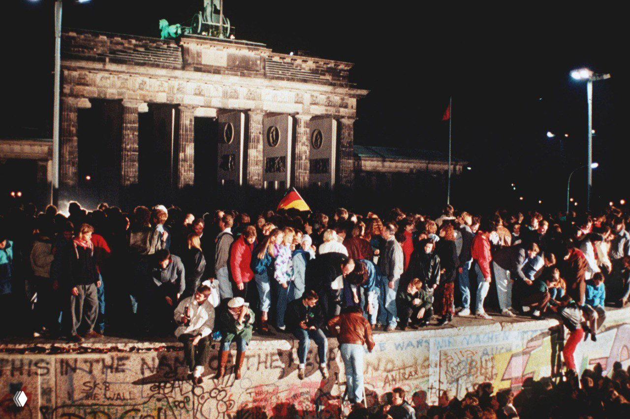 Толпа людей на верхней части Берлинской стены на фоне Бранденбургских ворот ночью, люди стоят и наблюдают, флаги и лампы освещают сцену исторического события.