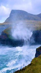 Прибрежные утёсы Фарерских островов, омываемые океанскими волнами; зелёные склоны и туманные горы на заднем плане.