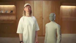 Человек демонстрирует рост и пропорции NEO на презентации: робот в вязаном 'костюме' стоит рядом с представителем компании, видно соотношение размеров.