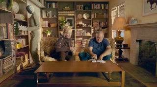 Сцена с пожилой парой и роботом NEO в домашней библиотеке: робот в мягкой одежде стоит возле полок, уютная обстановка и семейная сцена.