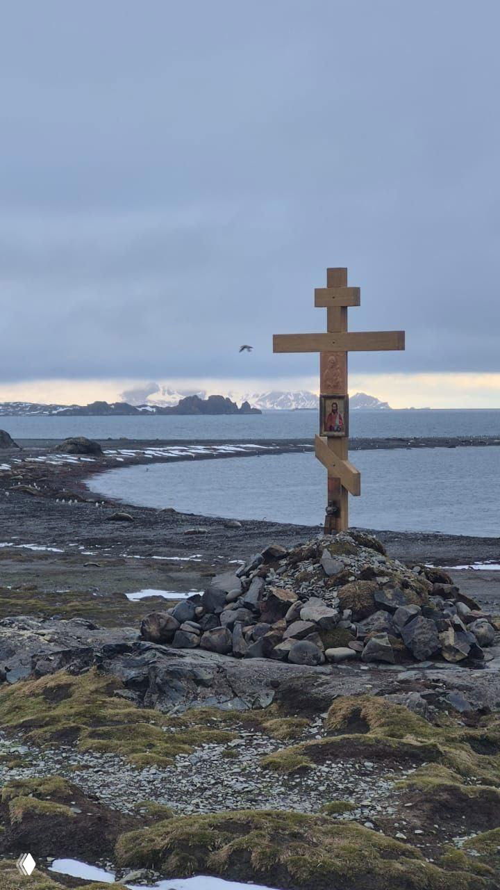 Деревянный православный крест на берегу острова Смоленск: икона на кресте, камни у подножия и антарктический морской пейзаж с дальними снежными горами.