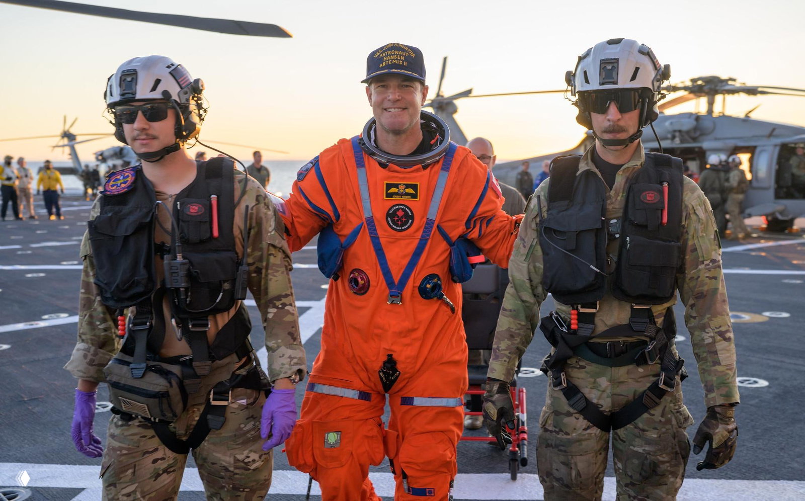 Астронавт идёт по палубе между военнослужащими в защитной экипировке; на заднем плане видны вертолёты и вечернее небо над океаном.