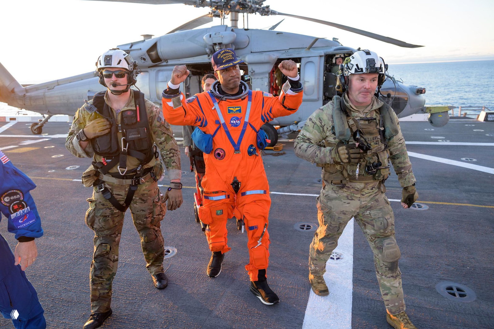 Астронавт идёт по палубе авианосца, поднимая руки в жесте радости, сопровождаемый спасателями в камуфляже — кадр возвращения миссии.