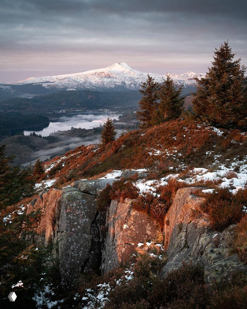 Широкий шотландский пейзаж: горы, озеро и хвойные деревья при тёплом утреннем свете, рельефное сочетание скал и растительности