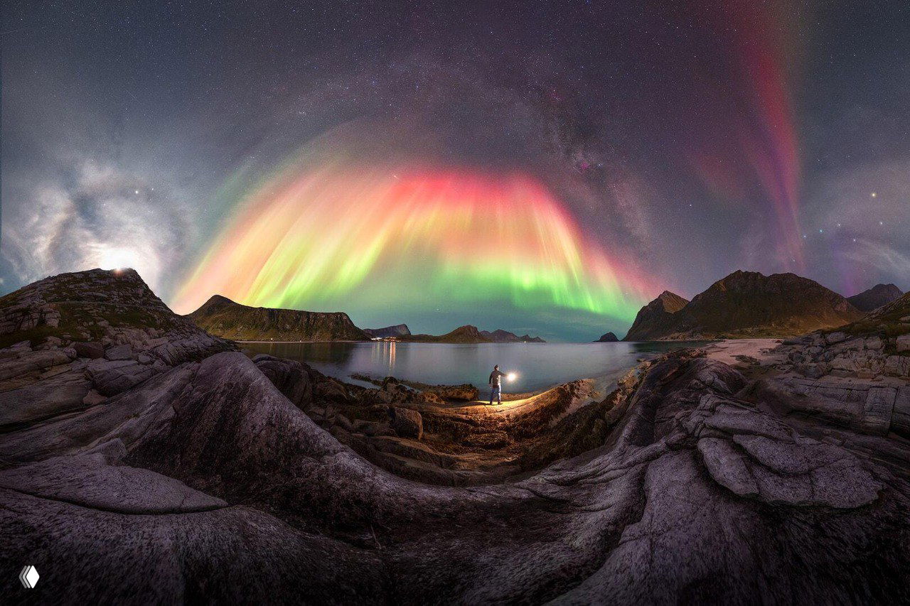 Панорамный вид бухты и скал с яркой зелено‑розовой аркой северного сияния над морским горизонтом и фотографом у кромки воды.