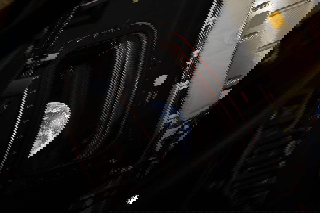 Вид на Землю через иллюминатор корабля: фрагмент диска планеты виден в проеме окна на фоне элементов внутренней обшивки.