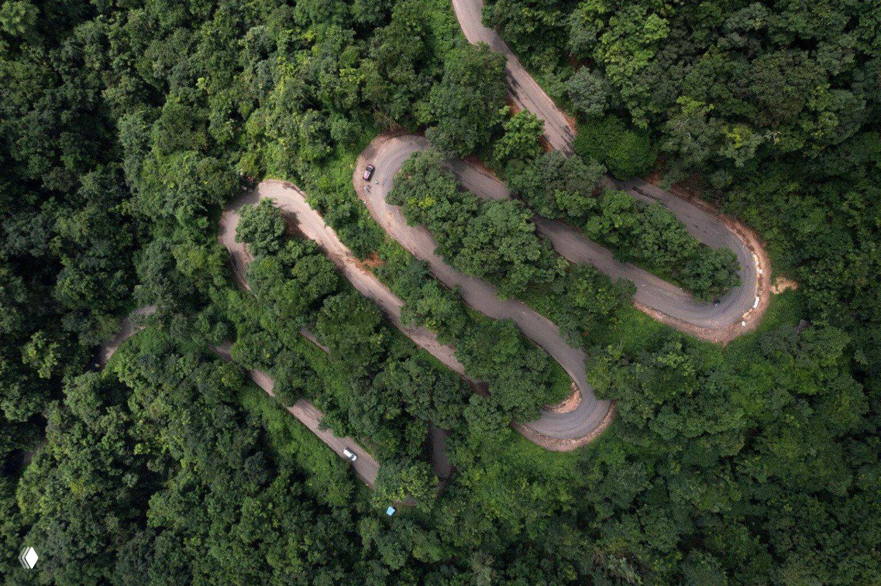 Аэрофотосъёмка извилистой горной дороги, петляющей сквозь плотный зелёный лес; вид сверху подчёркивает ритм изгибов и масштабы растительности.