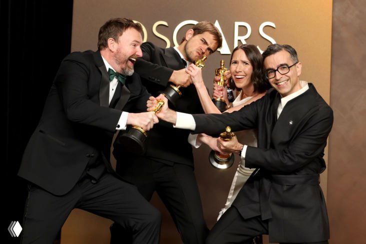 Группа людей в официальной одежде под позолоченным фоном держит статуэтки и демонстрирует радость победы на фотозоне «Оскар».