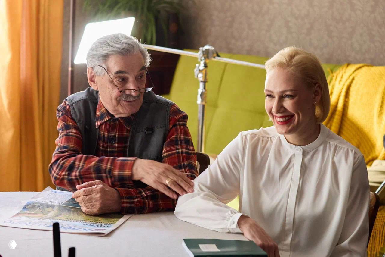 Сцена со съёмочной площадки: пожилой мужчина и женщина в светлой комнате улыбаются во время съёмки, заметно осветительное оборудование и реквизит.