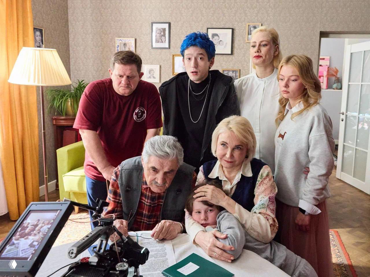 Группа актёров на съёмочной площадке: несколько персонажей разного возраста вокруг стола, камера, осветительные приборы и рабочая атмосфера съёмок.