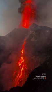 Ночное фото извержения вулкана Этна: яркая струя лавы, стекающая по склону, и плотный столб тёмного дыма над кратером.