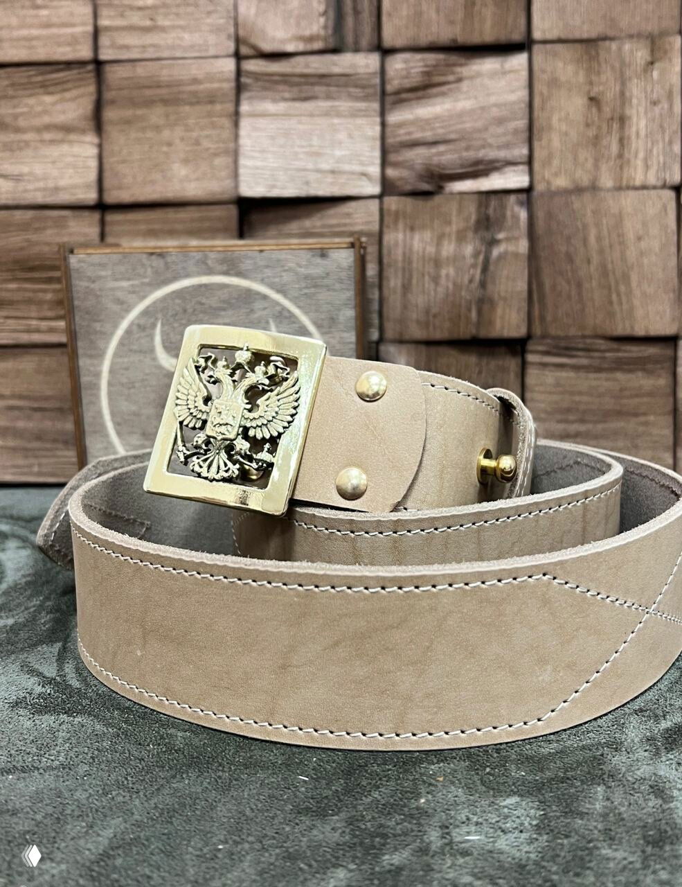 Светло-бежевый ремень из натуральной кожи с латунной пряжкой-гербом России, аккуратные строчки и видимая толщина заготовки ремня.