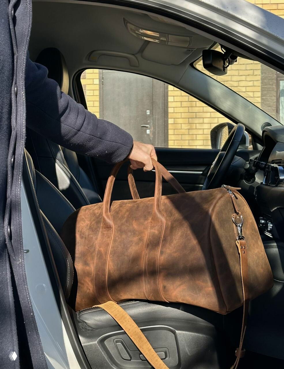 Коричневая сумка из состаренной кожи, человек переносит сумку в салон автомобиля, видно текстуру и форму дорожного саквояжа.