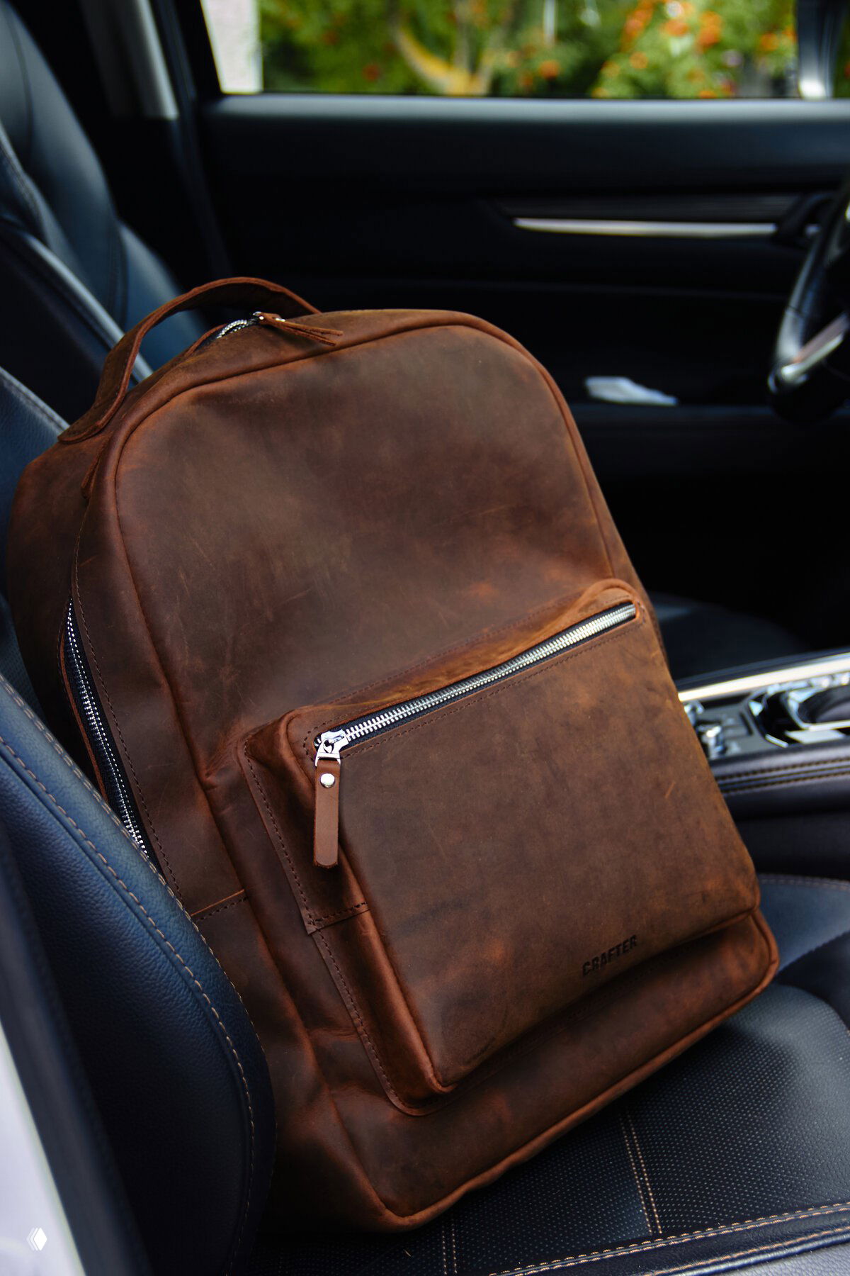 Тёмно-коричневый кожаный рюкзак на переднем сиденье автомобиля, виден внешний карман с молнией и естественная патина кожи — фото крупным планом.