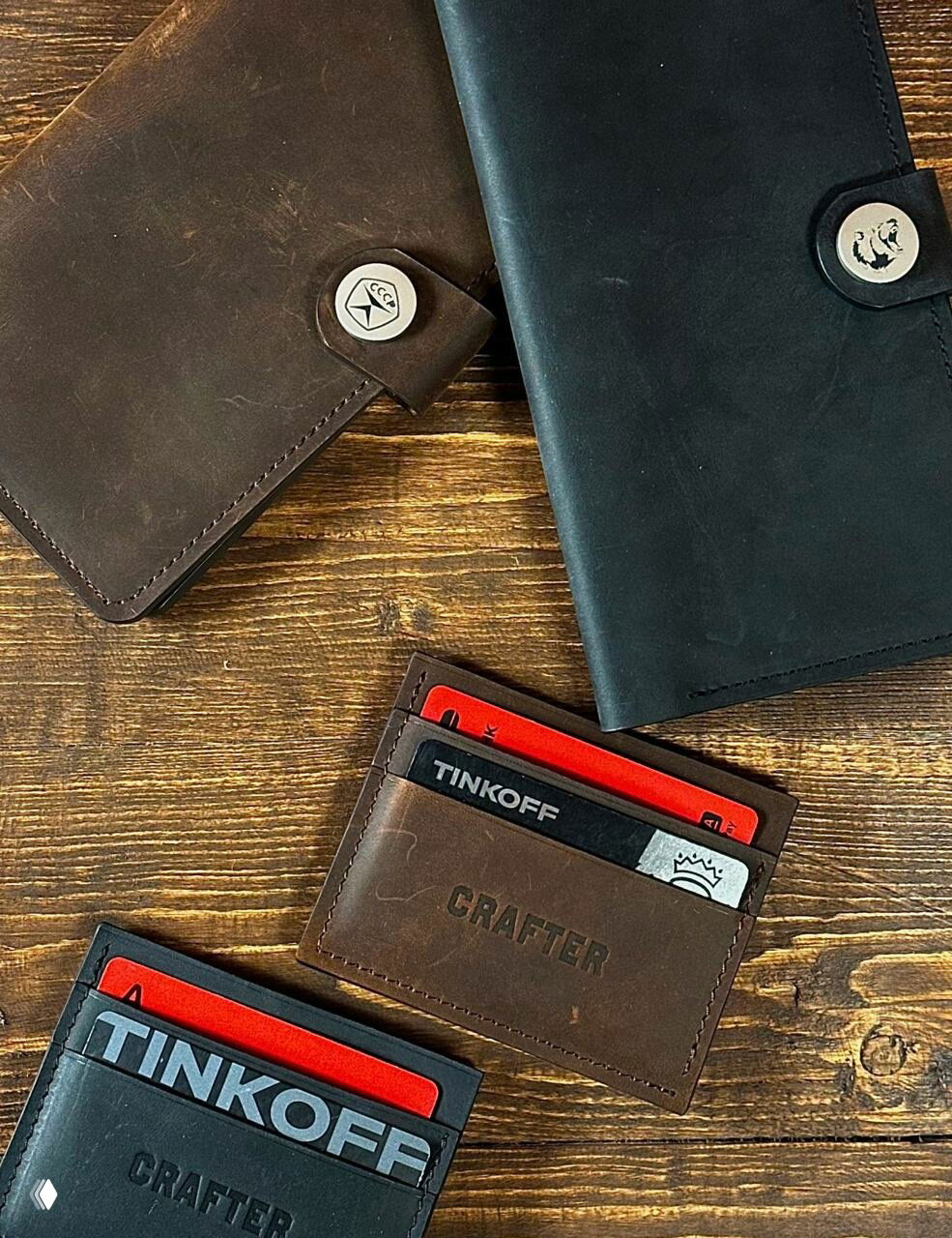 Несколько картхолдеров и портмоне из коричневой и чёрной кожи на деревянной поверхности, в одном держателе видна карта Тинькофф и тиснение бренда Crafter.