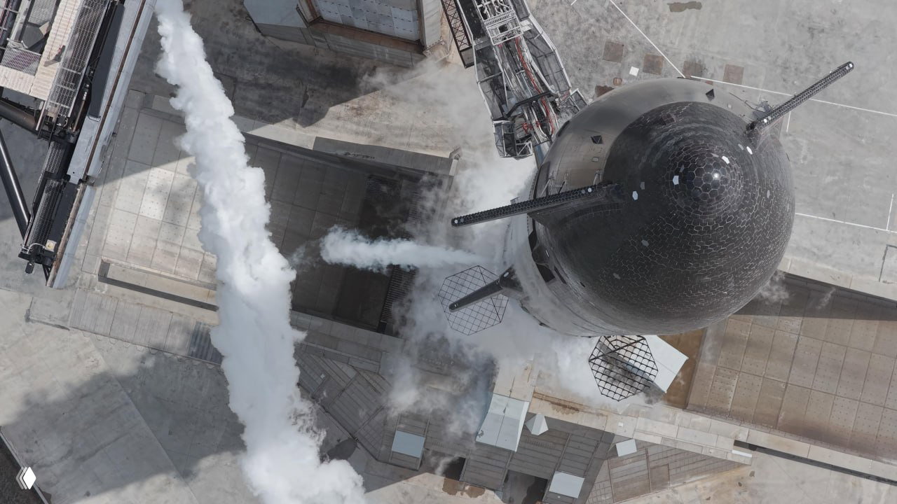 Вид сверху на носовую часть Starship и пусковую площадку: чёрная головная часть корабля, струи пара и элементы старта под углом.