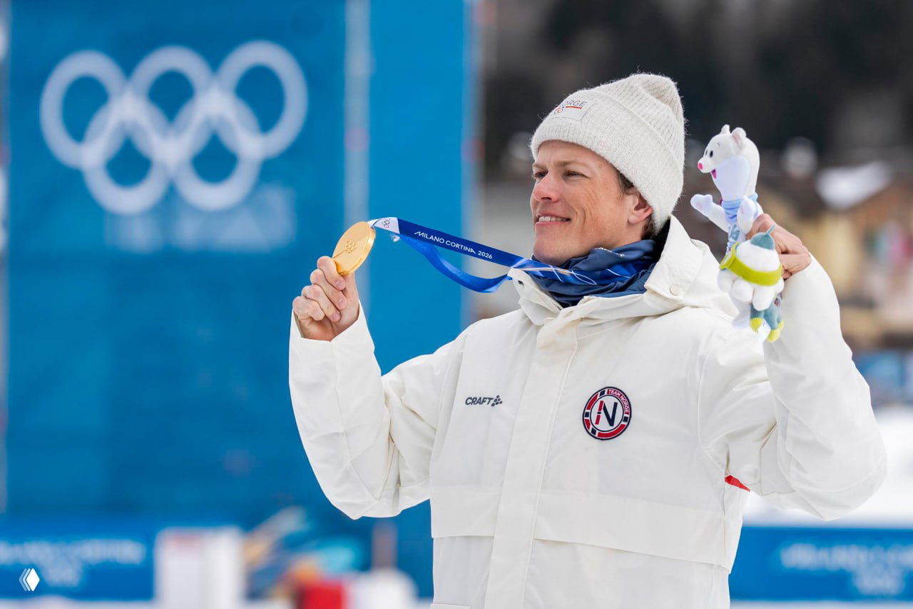 Йоханнес Клебо держит золотую медаль и талисман, улыбается на фоне синих элементов оформления Олимпиады и стартовой зоны в Кортине‑д’Ампеццо.