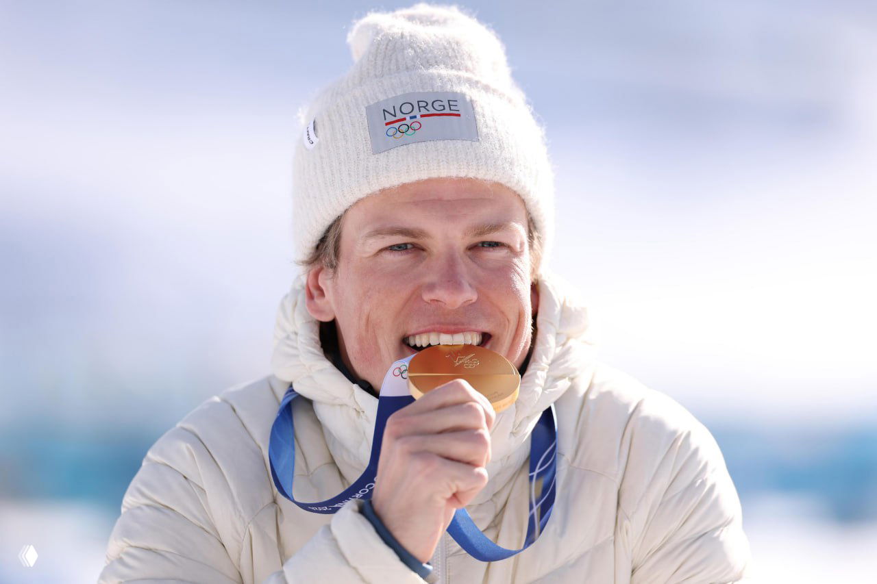 Крупный план Йоханнеса Клебо, который кусает золотую медаль в белой шапке с эмблемой NORGE на фоне снега и гор на Олимпийских играх 2026.