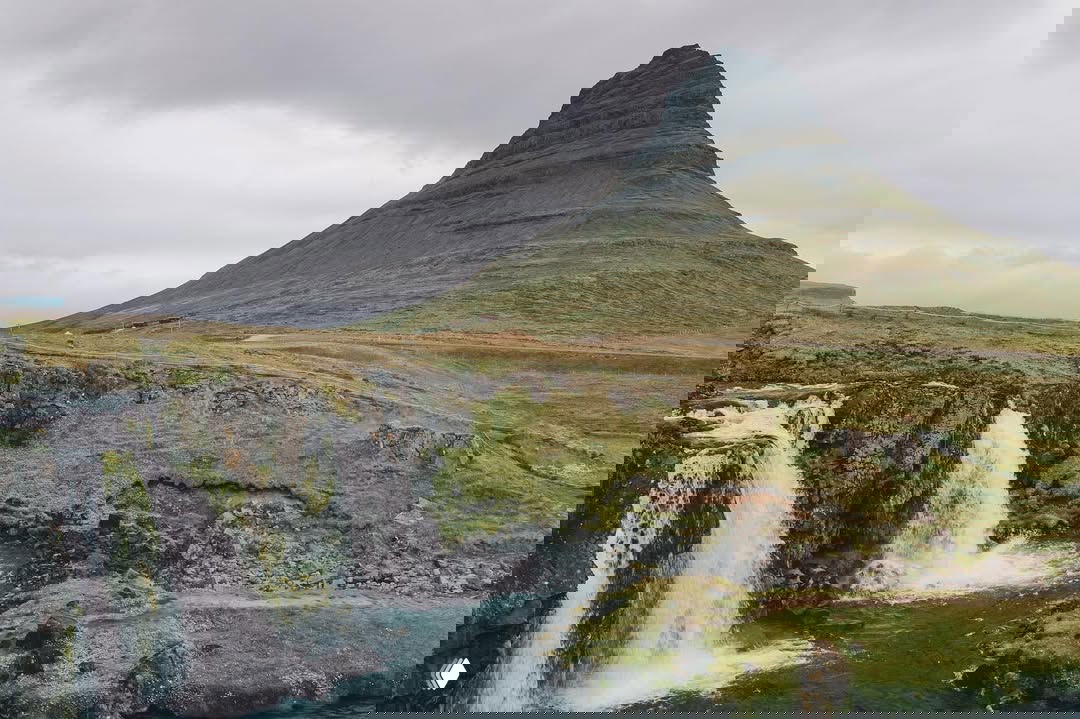 Вид на гору Киркьюфелл и трёхкаскадный водопад Киркьюфеллсфосс: бурные потоки воды на фоне зелёных скал и характерной конусообразной вершины.