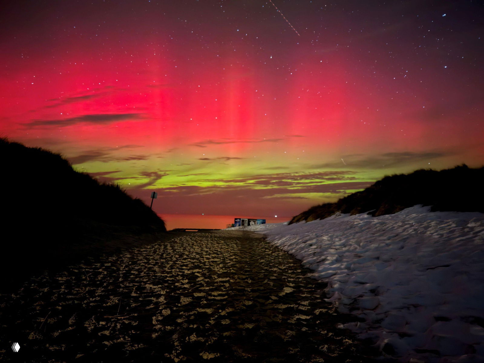 Широкая зелёная лента сияния над заснеженным берегом; на переднем плане видна тропинка и следы на снегу, ведущие к горизонту моря.