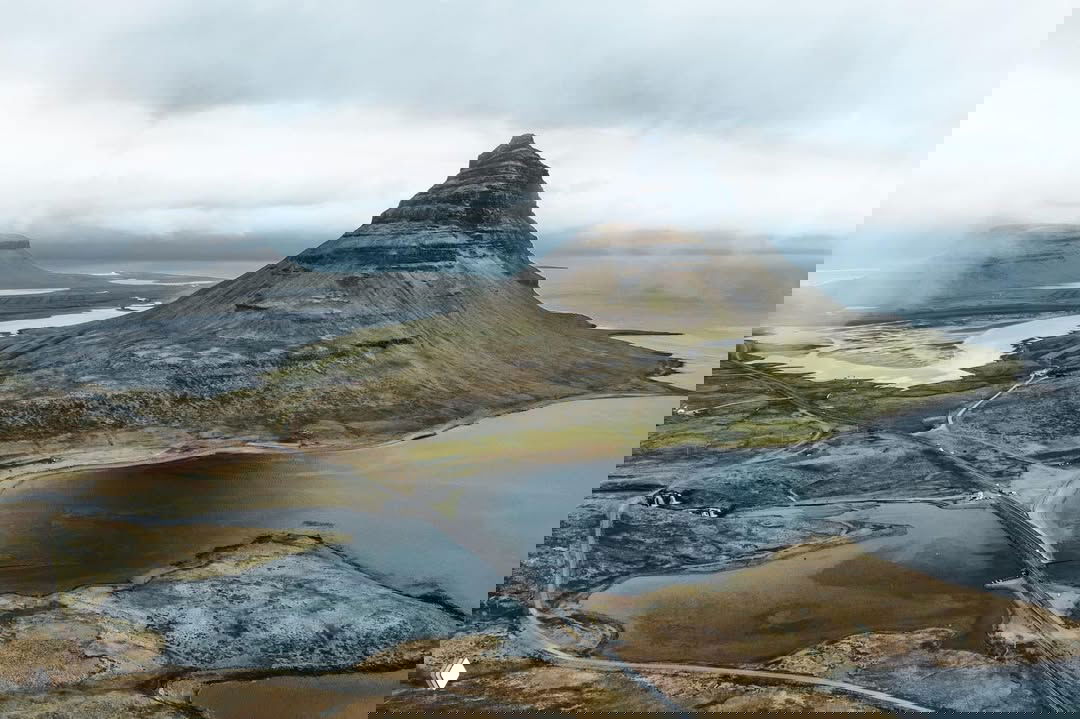 Аэрофотосъёмка Киркьюфелл: возвышающийся конус горы, окружающие лагуны, извилистые дороги и прибрежные участки полуострова Снайфедльснес в пасмурную погоду.