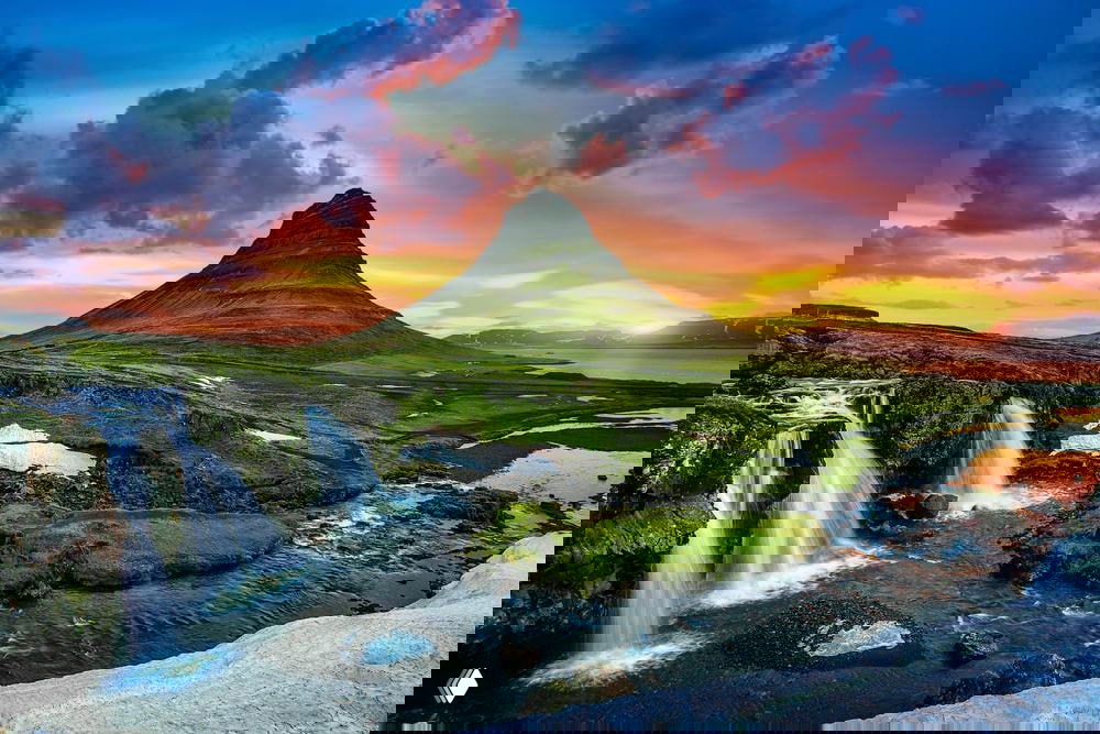 Панорамный вид на Киркьюфелл и водопад при ярком заходе солнца: драматические облака, отражения в воде и насыщенная зелёная растительность побережья.