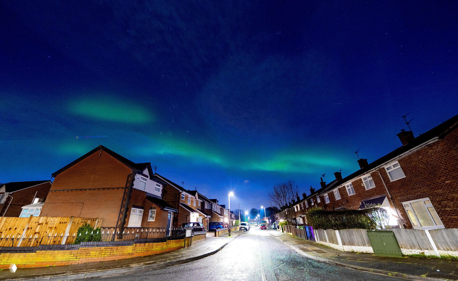 Интенсивное зелёное северное сияние над рядом деревянных домов в снегу; в одном доме горят окна, создавая контраст тёплого света и зелёного неба.
