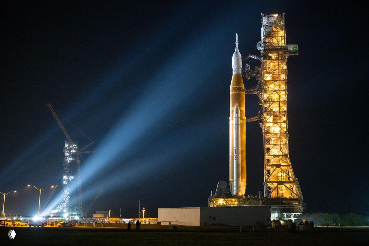 Ракета-носитель SLS у подсвеченной стартовой мачты ночью, лучи прожекторов освещают силуэт ракеты — атмосферный кадр подготовки к пуску.