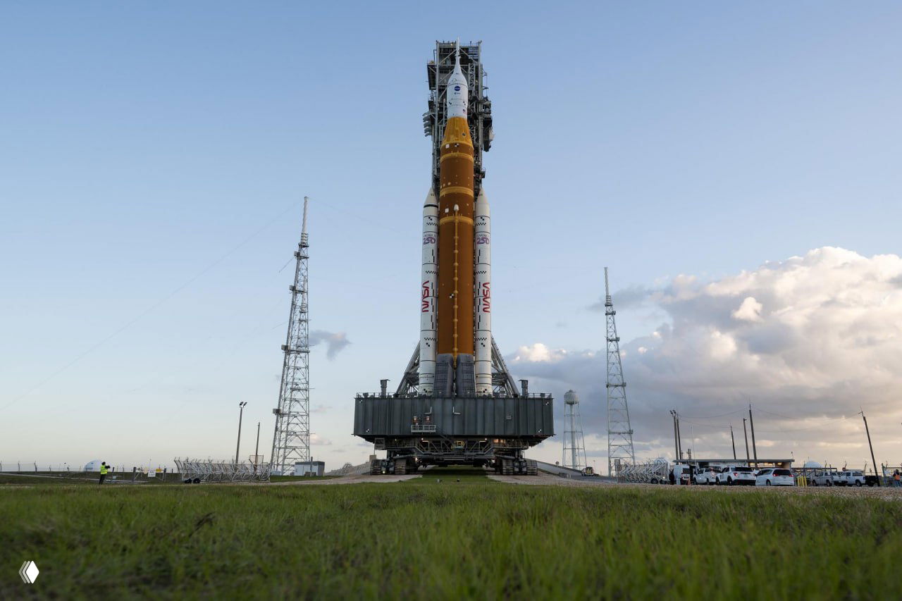 Ракета SLS на транспортной платформе движется по траектории к стартовой площадке, вид с уровня грунта и неба с облаками — этап вывозки.