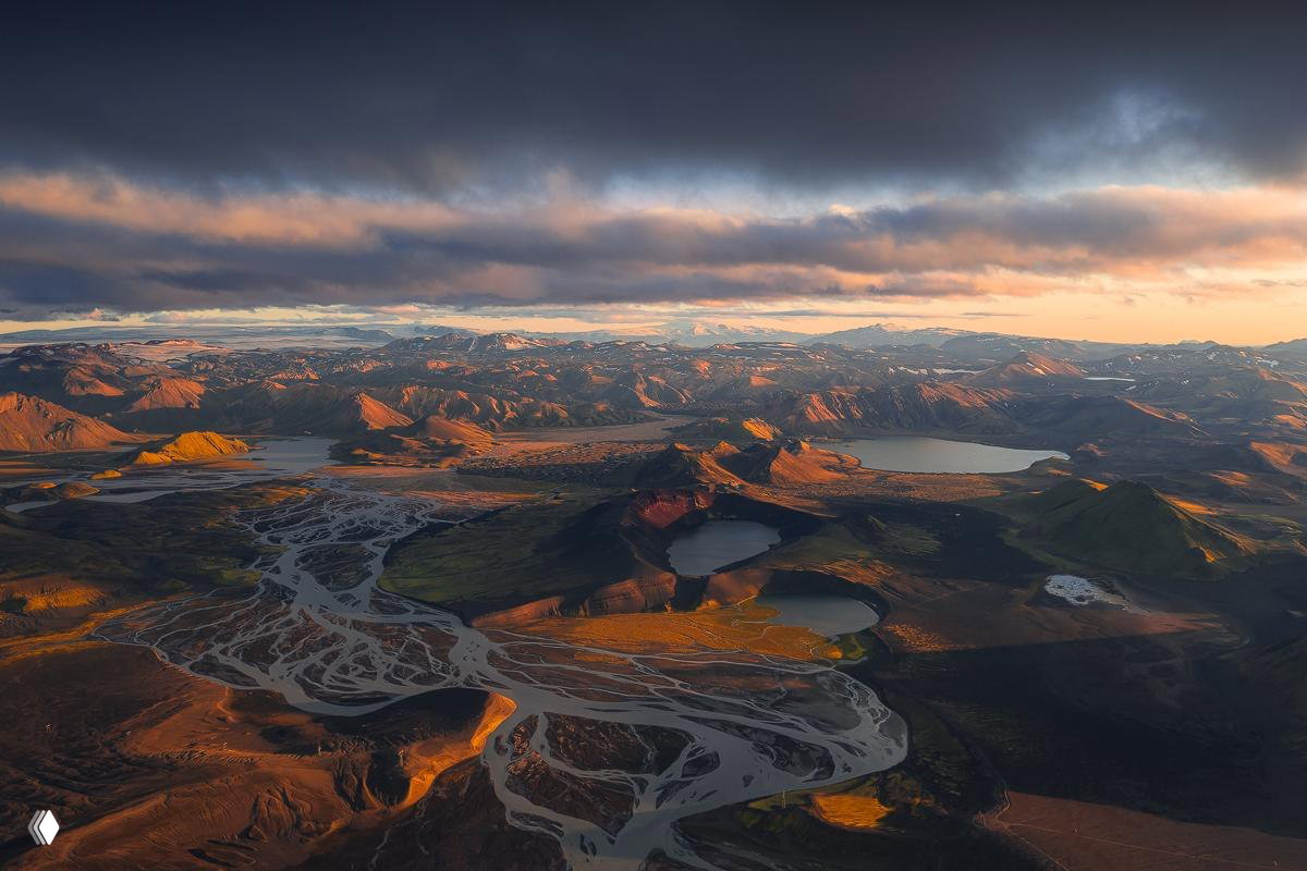 Аэрофотосъёмка обширного лавового и озёрного ландшафта, извилистые русла рек и тихие озёра под тёмным небом на рассвете.