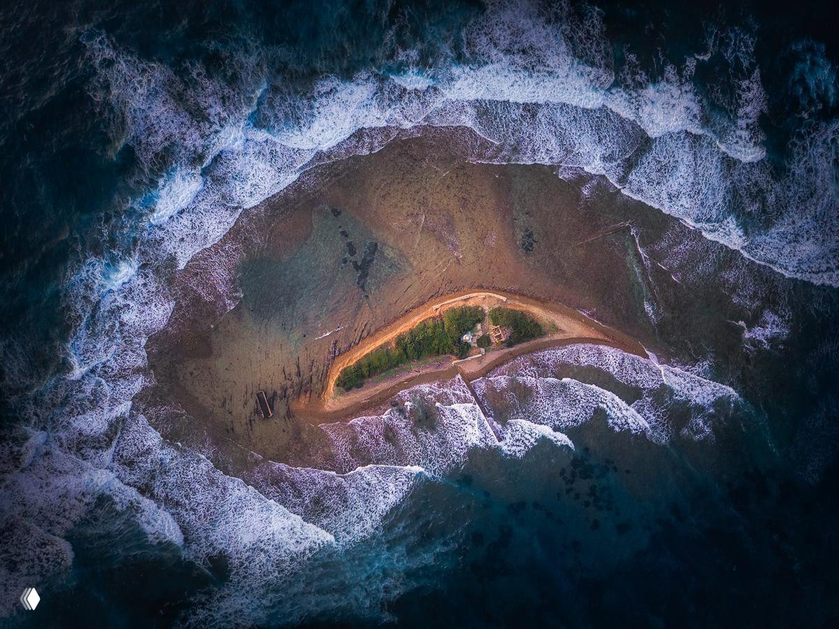 Вид сверху на узкий островок с пляжем и растительностью, окружённый пенящимися волнами и контрастными морскими оттенками.