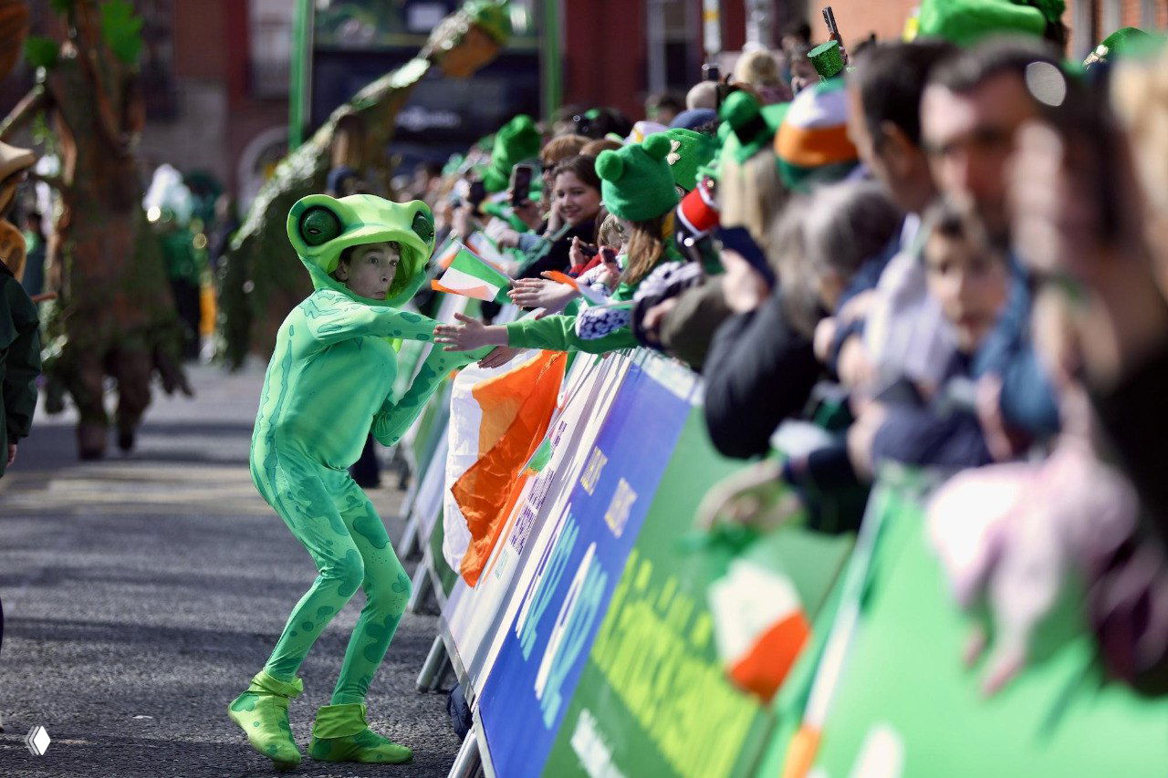 Ребёнок в костюме лягушки протягивает руку к публике вдоль ограждения парада в Дублине; флаги Ирландии и зрители на заднем плане.