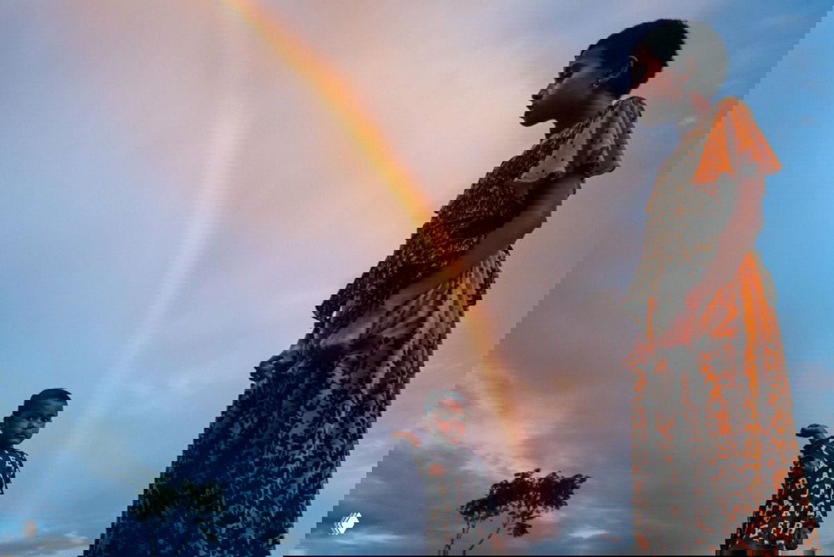 Два ребёнка на фоне яркой радуги в небе; композиция подчёркивает контраст цветной одежды и безмятежный пейзаж с низким горизонтом.