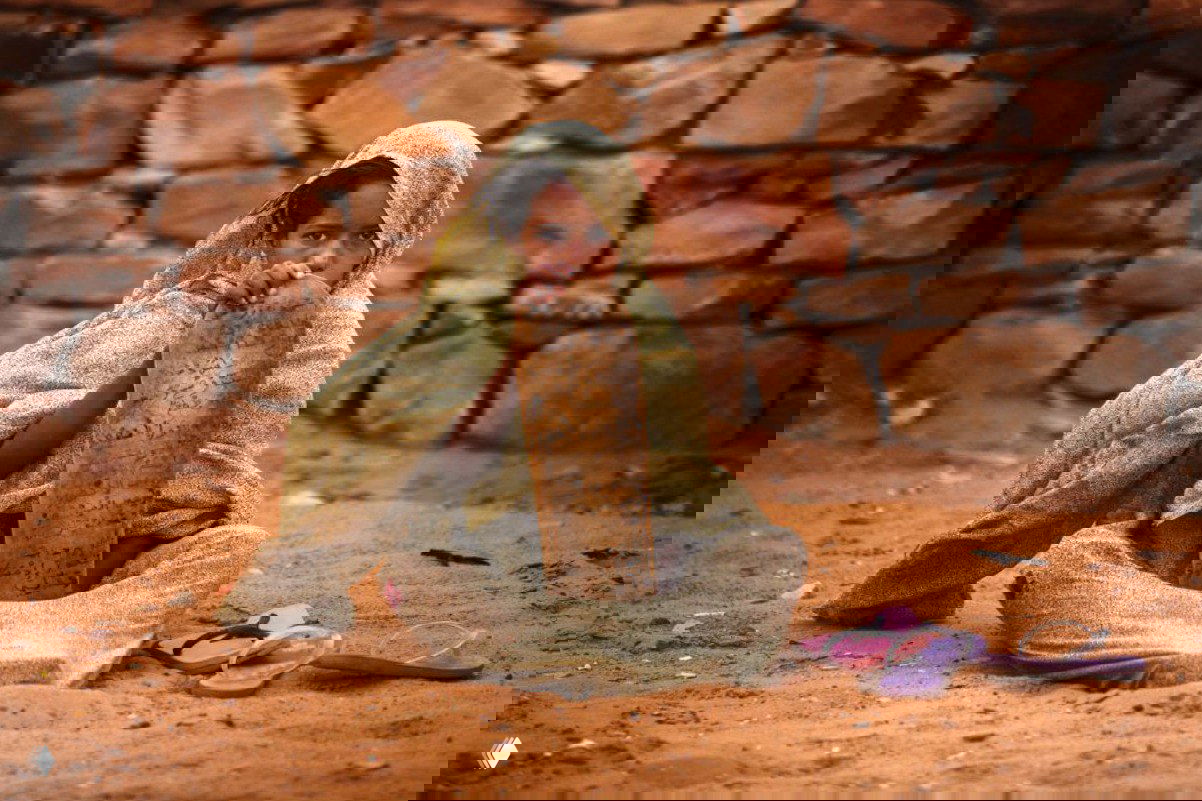 Ребёнок, обёрнутый в ткань, сидит на песке и держит деревянную дощечку с надписями; сцена передаёт уязвимость и культурный контекст места.