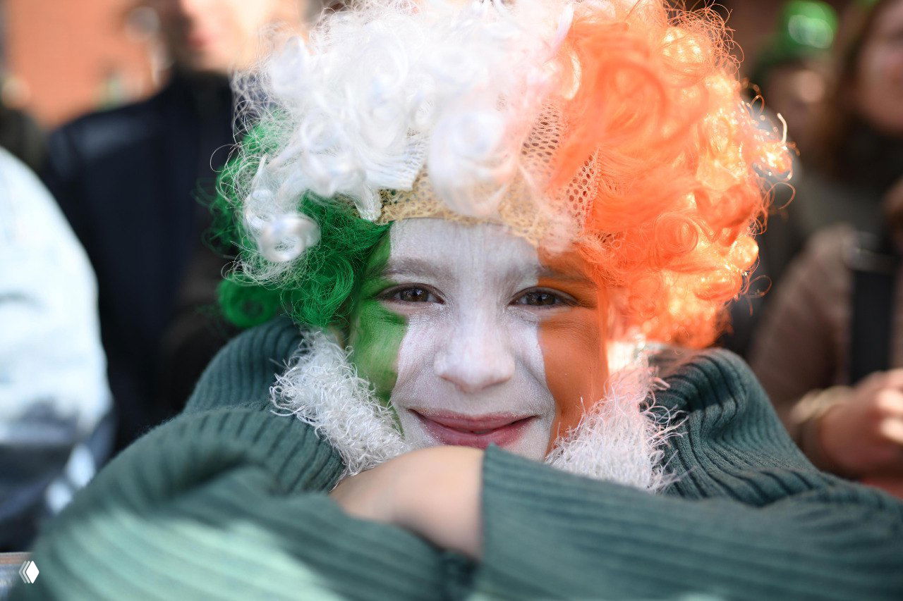 Крупный план ребёнка с раскрашенным лицом в цветах ирландского флага и париком на параде в Дублине, улыбающаяся публика за кадром.