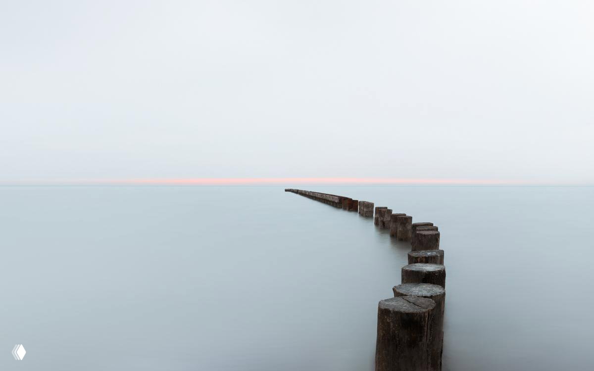 Минималистичный морской пейзаж с рядом деревянных сваёз, уходящих в гладь спокойного моря и тонкой светлой полосой горизонта.