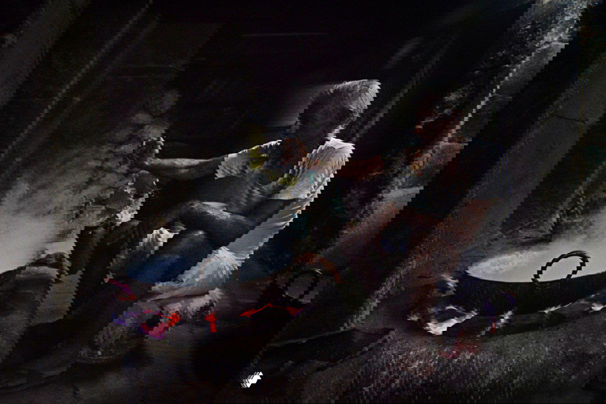 Мужчина у большого котла на открытом огне в тёмном помещении; пар, фактура стен и огонь создают атмосферу традиционного приготовления пищи.