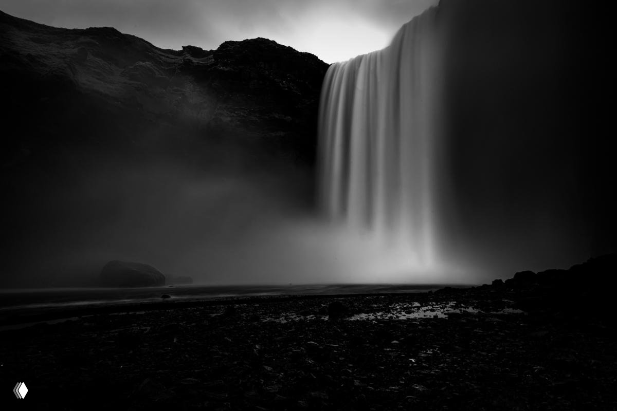 Мощный водопад в монохромной обработке: струи воды стекают по скале, туман и контрастная композиция с каменистым берегом.