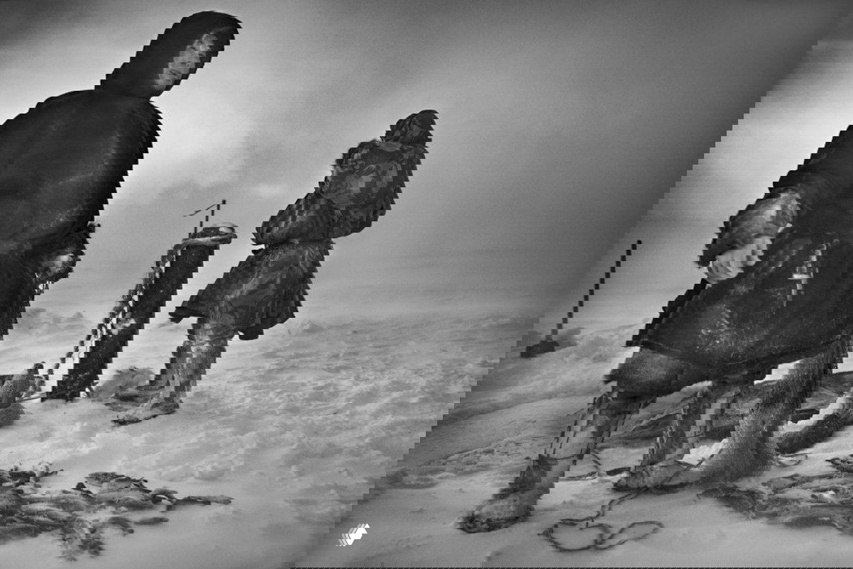 Чёрно-белый документальный снимок людей в тяжёлой одежде на заснеженной равнине; внимание к деталям экипировки, погодным условиям и композиции.