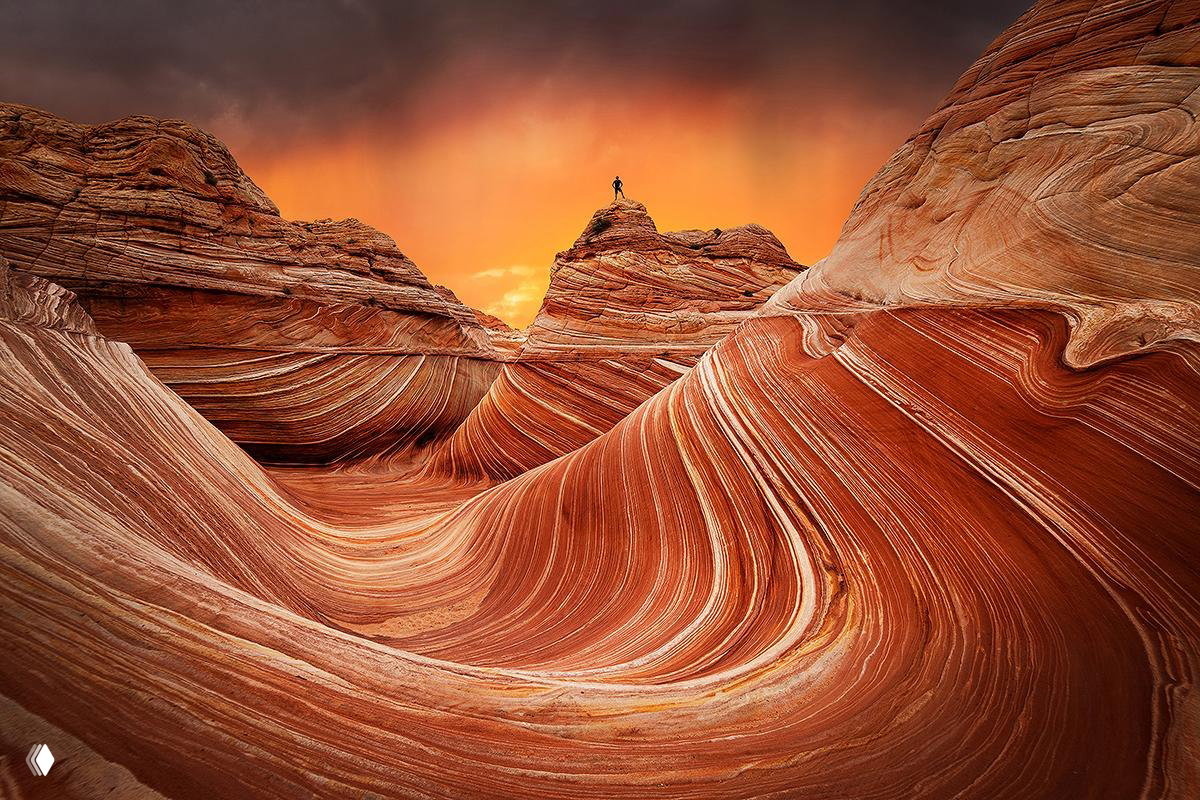 Извилистые слоистые песчаные образования и волнообразный рельеф в тёплых тонах, одиночный силуэт человека сверху у вершины утёса.