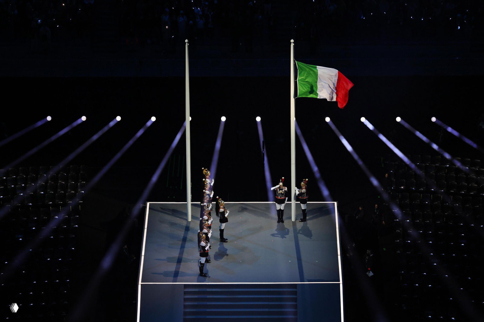 Почётный караул и поднятие итальянского флага на сцене стадиона, яркие лучи прожекторов и тёмный фон трибун — момент церемонии в Милане.
