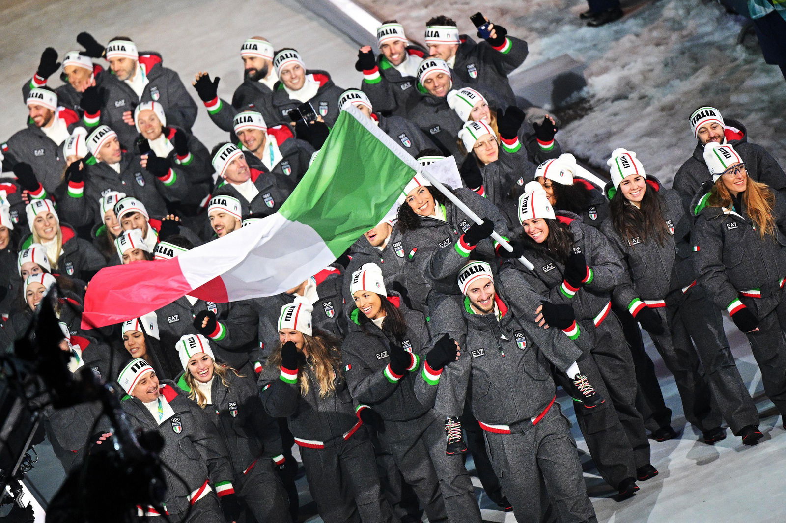 Спортсмены итальянской сборной в фирменной форме и шапках с надписью ITALIA идут, поднимая итальянский флаг в процессе парада на открытии Игр.