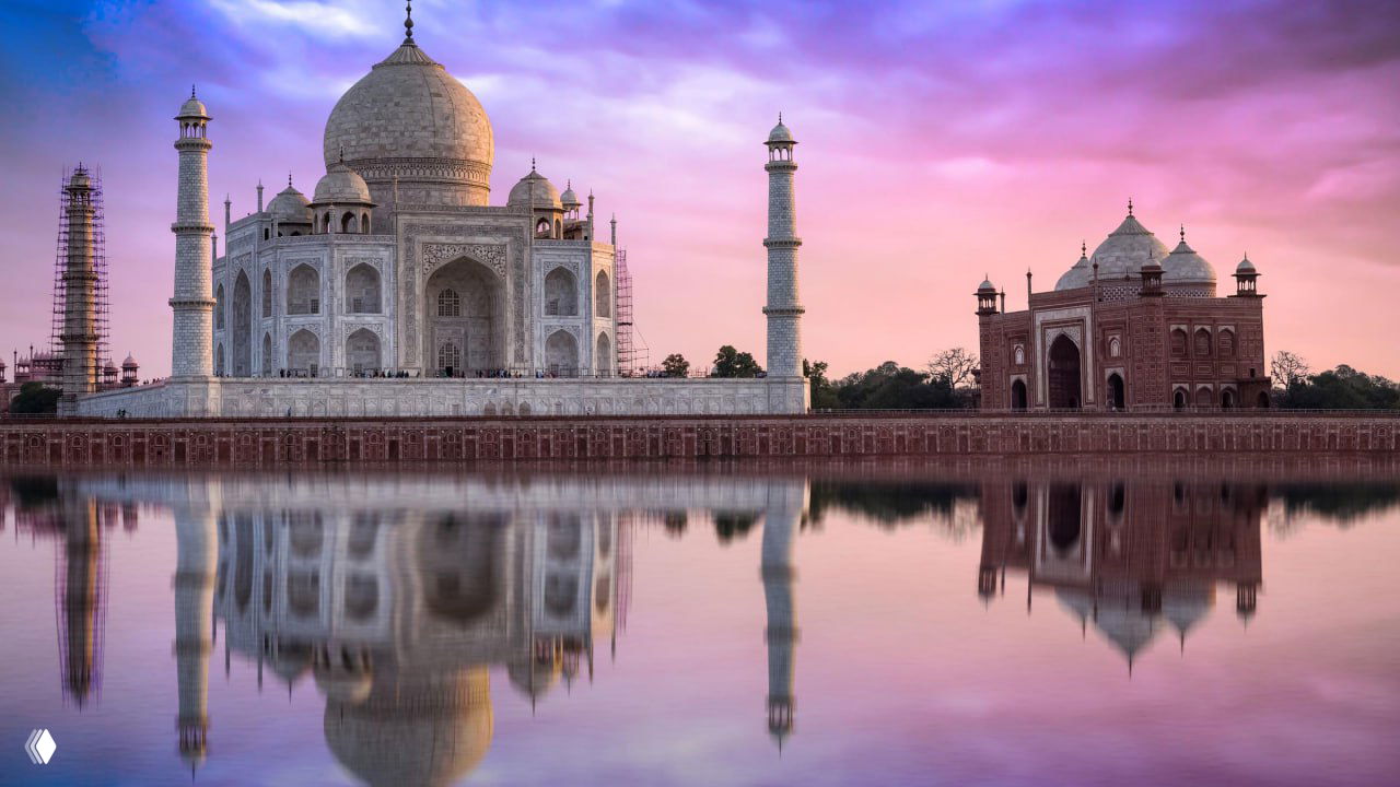 Тадж‑Махал и его зеркальное отражение в спокойной воде при цветном небе, вид с противоположного берега с заметными минаретами и боковыми постройками.
