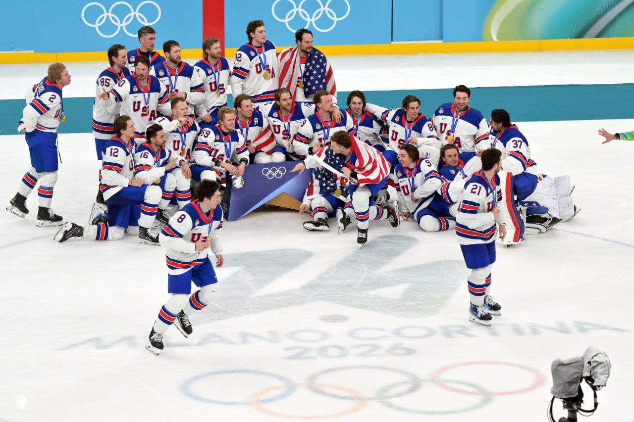 Команда сборной США на льду группой фотографируется с олимпийской символикой и золотыми медалями после победы в финале хоккейного турнира ОИ‑2026 в Милане.