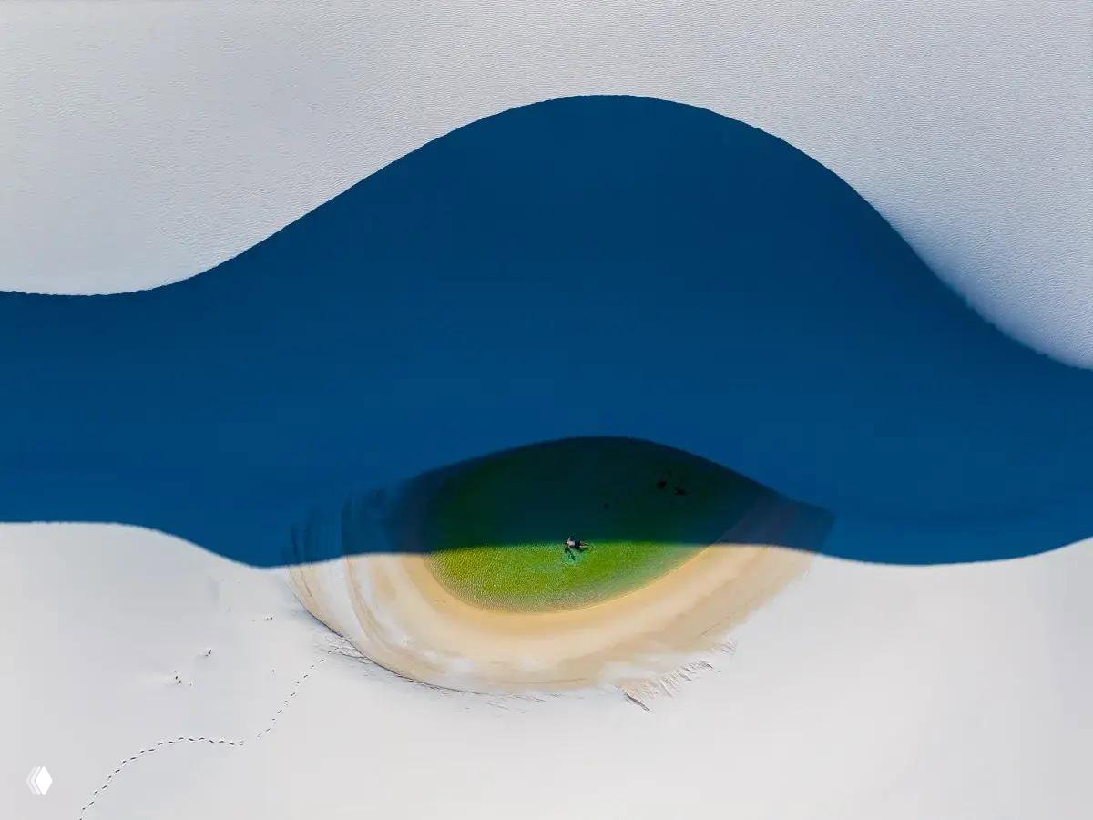 Минималистичный аэрофрейм с островком и пляжем: глубокая синяя полоса воды, зелёное пятно травы и белый песок, снято сверху дроном.