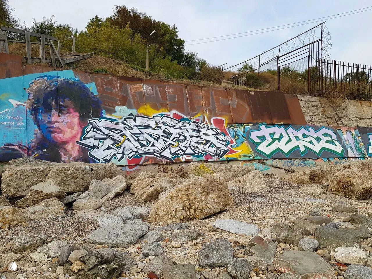 Граффити на береговом укреплении среди камней и обломков, цветные уличные росписи на фоне рабочего приморского ландшафта.