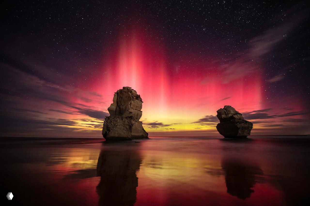 Ночное небо с ярким зелёным сиянием и образом кометы в левой части кадра, отражающимся в спокойной водной поверхности на переднем плане.